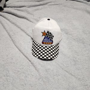 May include: White baseball cap with a black and white checkered brim. The front of the cap has a patch with a star and the text "All-American Soap Box Derby 2000".