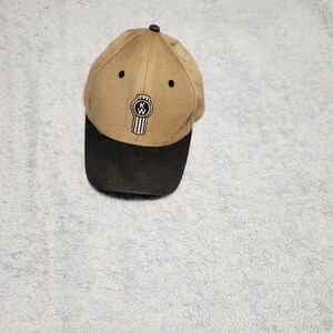 May include: A tan and black baseball cap with a black and white Kenworth logo embroidered on the front.