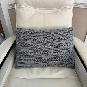 May include: A rectangular, gray crochet pillow with a textured, openwork design. The pillow is placed on a white leather chair. The crochet pattern features evenly spaced holes, adding visual interest to the home decor item.