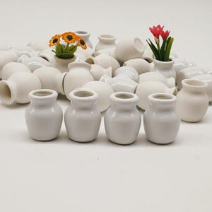 Könnte beinhalten: Eine Ansammlung kleiner, weißer Keramikvasen, einige mit künstlichen Blumen in Orange und Rot. Die Vasen haben verschiedene Formen und Größen mit einer glatten, matten Oberfläche. Das Thema deutet auf ein Bastel- oder Dekorationsartikel hin.