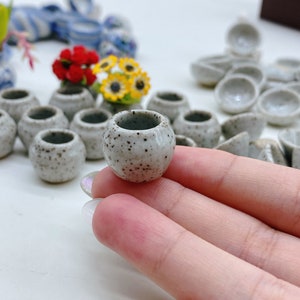 May include: A collection of small, speckled ceramic vases. The vases are a light gray color with dark brown speckles. Some vases hold miniature flowers. The vases are round with a wide opening. The image is taken from a close-up angle.