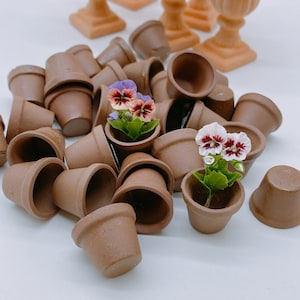 May include: A collection of small brown terracotta pots with two miniature potted plants featuring purple and white flowers. The pots are arranged in a pile on a white surface.