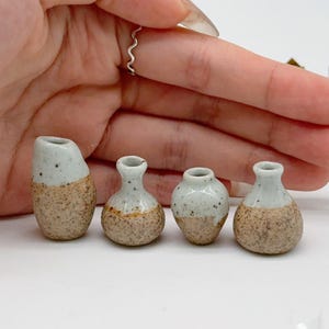 Puede incluir: Cuatro jarrones de cerámica en miniatura. Cada jarrón tiene una base moteada de color marrón claro y una parte superior gris claro. Los jarrones varían en forma, uno es más angular y los otros redondos. Los jarrones son pequeños, probablemente para fines decorativos.