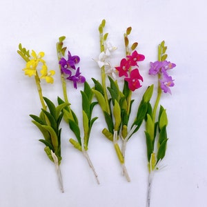 May include: Five artificial orchid flower stems with green leaves. The flowers are yellow, pink, purple, and white.