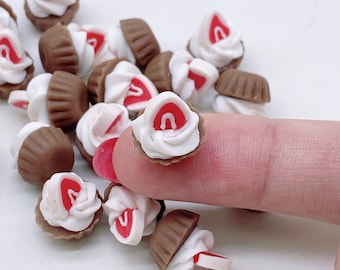 May include: Miniature brown cupcake charms with white frosting and a red strawberry slice on top. These small, detailed charms are perfect for crafting, jewelry making, or other creative projects. The cupcakes are arranged on a white surface.