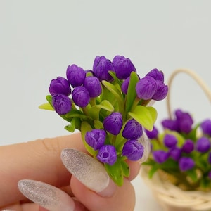 May include: A miniature bouquet of purple tulips with green leaves. The small flowers are held in a hand, with a wicker basket of similar flowers in the background. The tulips are a vibrant purple color.