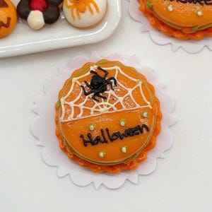 Pode incluir: Um bolo laranja em miniatura decorado com uma teia de aranha branca e uma aranha preta. O bolo está sobre um papel branco e diz "Halloween" em glacê preto.