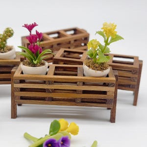 May include: A miniature wooden planter with two compartments, each holding a potted plant. The plant on the left has pink flowers and green leaves, while the plant on the right has yellow flowers and green leaves.