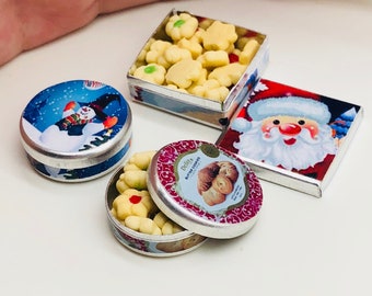 May include: Three miniature tin boxes filled with cookies. One box has a snowman design, another has a Santa Claus design, and the third has a red and white design with the text "Dello's Butter Cookies".