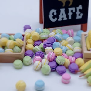 Può includere: Una varietà di macarons colorati in tonalità pastello di rosa, viola, verde, giallo e blu. I macarons sono disposti in un mucchio su una superficie bianca. La parola "CAFE" è visibile su un cartello nero sullo sfondo.