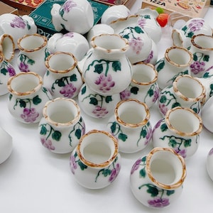 Puede incluir: Una colección de tazas de té de cerámica blanca con un diseño floral con flores moradas y hojas verdes. Las tazas están dispuestas en un patrón aleatorio sobre una superficie blanca.