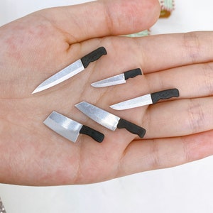 Puede incluir: Cinco cuchillos en miniatura con mango negro y hojas plateadas en una mano. Los cuchillos son de diferentes tamaños y formas.