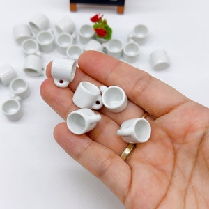 Może przedstawiać: Ręka trzymająca kolekcję małych białych ceramicznych kubków. Kubki są wszystkie tego samego rozmiaru i kształtu, a ich design jest prosty i klasyczny.
