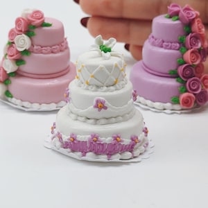 Puede incluir: Mini pastelitos decorativos en rosa, blanco y morado. Los pasteles son de varios niveles y están adornados con decoraciones florales. Un pastel es blanco con un diseño de enrejado, y otro es rosa con rosas blancas y rosas.