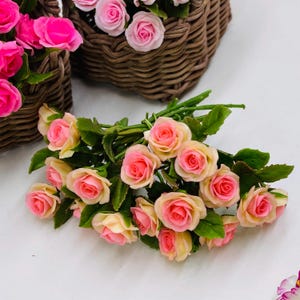 以下が含まれることがあります： 淡いピンクの中心部とクリーム色の外側の花びらを持つ造花のバラの束で、緑の葉が添えられています。バラは房状に配置されており、背景にはバラで満たされた茶色の編み込みバスケットがいくつか見えています。