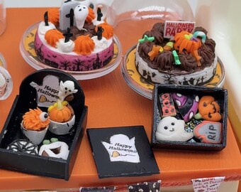 Könnte beinhalten: Eine Sammlung von Miniatur-Desserts zum Thema Halloween, darunter zwei Kuchen auf Ständern, zwei schwarze Schachteln mit kleinen Leckereien in Form von Geistern, Kürbissen und Grabsteinen sowie ein Schild mit der Aufschrift Happy Halloween.