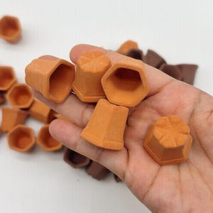 Può includere: Una collezione di piccoli vasi da fiori in miniatura color terracotta. I vasi hanno una forma esagonale e una superficie strutturata. Alcuni sono tenuti in mano, mentre altri sono sparsi su una superficie bianca. Ideali per piccole piante o lavori artigianali.