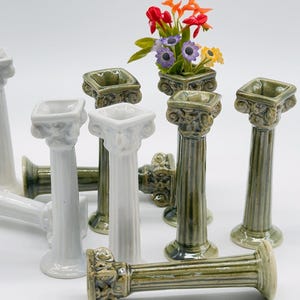 Könnte beinhalten: Ein Set aus Miniatur-Säulen aus Keramik in weißer und grüner Glasur. Einige der Säulen sind mit einer quadratischen Öffnung versehen, während andere eine runde Öffnung haben. Ein kleiner Blumenstrauß wird in einer der Säulen ausgestellt.