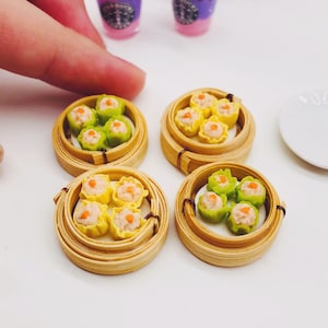 Op de afbeelding: Vier miniatuur bamboe stoommandjes gevuld met kleine, gestoomde dumplings. De dumplings zijn geel met groene toppen en hebben een klein oranje midden. De mandjes zijn gerangschikt op een wit oppervlak.