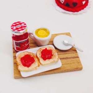 Puede incluir: Un juego de desayuno en miniatura en una bandeja de madera con dos rebanadas de tostadas con mermelada roja, un pequeño frasco de mermelada de fresa Smucker's, una taza de líquido amarillo y un pequeño plato blanco con una cuchara.