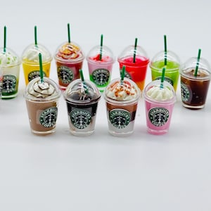 Op de afbeelding: Een set van tien miniatuur Starbucks-koffiebekers met verschillende toppings en kleuren, waaronder chocolade, karamel, slagroom en fruit. De bekers zijn gemaakt van plastic en hebben het Starbucks-logo erop.