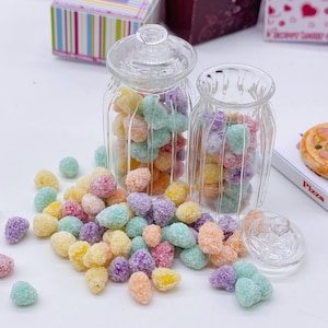 Puede incluir: Dos tarros de vidrio transparente llenos de caramelos coloridos. Los caramelos son pequeños y redondos, y están cubiertos con una capa de azúcar blanca. Los tarros están sobre una superficie blanca, y hay algunos caramelos esparcidos alrededor.