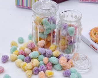 May include: Two clear glass jars filled with colorful candy. The candy is small and round, and it is covered in a white sugar coating. The jars are sitting on a white surface, and there are some candies scattered around them.