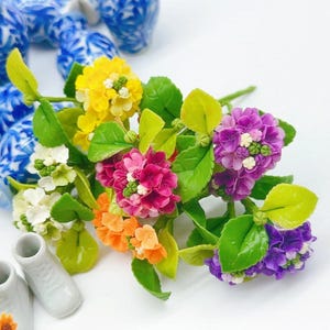 May include: A bouquet of colorful artificial flowers with green leaves. The flowers are made of polymer clay and are arranged in a cluster. The flowers are in shades of yellow, orange, pink, purple, and white.