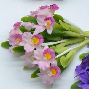 以下が含まれることがあります： 緑の葉が付いたピンクと白の蘭の花のブーケ。花はポリマークレイで作られており、リアルな外観をしています。