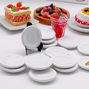 Könnte beinhalten: Eine Sammlung von Miniatur-Tellern aus weißer Keramik, von denen einer auf einem kleinen schwarzen Staffeleiständer präsentiert wird. Das Set besteht aus mehreren runden, glatten und schlichten Tellern, die in einer Gruppe angeordnet sind.