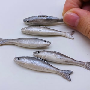 May include: A collection of small, silver-colored fish figurines. Each figurine has a detailed design, including fins and eyes. The fish are arranged on a white surface, with one being held by a hand.