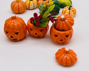 May include: A collection of ceramic Halloween-themed pumpkins in various sizes and styles. Some are carved with jack-o'-lantern faces, while others are plain. One pumpkin holds a small floral arrangement. The pumpkins are orange, green, and white.