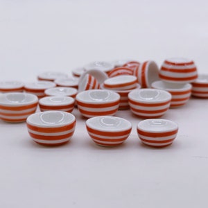Może przedstawiać: Zbiór miniaturowych białych ceramicznych miseczek z czerwonymi paskami. Miseczki są ułożone w stos i na białym tle.