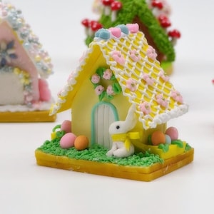 May include: A miniature yellow Easter house with a white door, a lattice roof adorned with pink flowers, and a white bunny with a yellow scarf. The house sits on a green base with colorful Easter eggs. Other decorative houses are in the background.