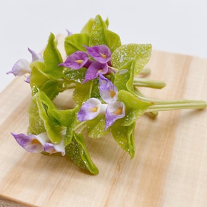 以下が含まれることがあります： 緑の葉が付いた紫と白の人工の花の房。花は花束にまとめられ、木製の表面の上に置かれています。