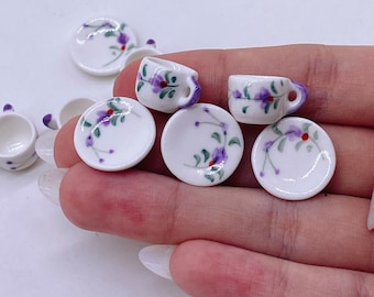 May include: A set of miniature teacups and saucers with a floral design. The cups and saucers are white with purple flowers and green leaves.