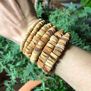 以下が含まれることがあります： 薄い茶色とベージュのセラミックビーズで作られた重ね付けブレスレットのセット。ブレスレットは手首に着用されています。