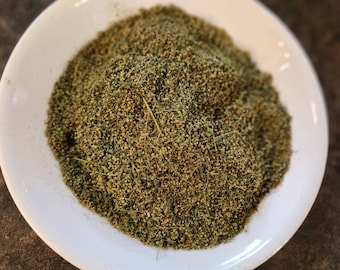 May include: A white ceramic plate filled with a mound of dried green herbs. The herbs have a fine, textured appearance with small, seed-like elements and a few strands of straw. The plate is set against a dark brown surface.
