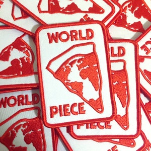 Puede incluir: Un parche blanco con costuras rojas presenta el texto "WORLD PIECE" en rojo. El parche representa un contorno rojo del mundo en forma de porción de pizza.