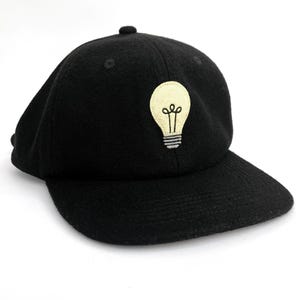 May include: Black baseball cap featuring a cream-coloured lightbulb patch with black detailing. The cap has a curved peak and a small button on top.
