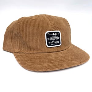 Peut inclure: Casquette de baseball en velours côtelé marron avec une visière incurvée. Un écusson carré affiche le texte "Thank Cod It's Friday" et un dessin d'arête de poisson. La casquette est de couleur neutre et convient à un usage quotidien.