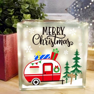 May include: A white light-up glass block with a red and white camper van, two green Christmas trees, and the text "Merry Christmas" in black. The camper van has a wreath on the back window and a sign that says "Welcome".