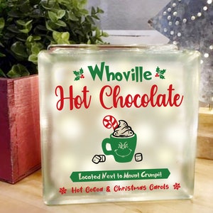 May include: A white glass block with a green and red design. The text on the block reads "Whoville Hot Chocolate" and "Located Next to Mount Crumpit Hot Cocoa & Christmas Carols".