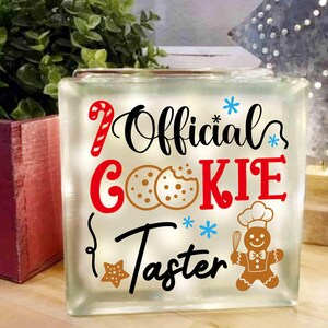 May include: A white glass block with the text "Official Cookie Taster" in black and red lettering. There are also images of gingerbread cookies and candy canes.