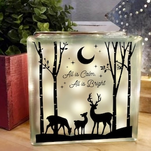 May include: A glass block with a white background and black silhouette images of trees, a crescent moon, and deer. The text "All is Calm, All is Bright" is printed on the block.