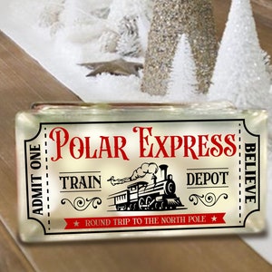 May include: A white ticket with red text that reads "ADMIT ONE" and "POLAR EXPRESS". The ticket has a black and white illustration of a train with the words "TRAIN" and "DEPOT" above and below it. The ticket also has a red star and the text "ROUND TRIP TO THE NORTH POLE" and "BELIEVE".