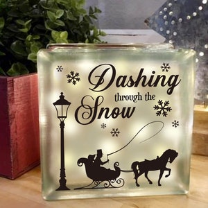 May include: A clear glass block with the words "Dashing through the Snow" in a decorative font. The block features a black silhouette of a horse-drawn sleigh, snowflakes, and a lamppost. The block is lit from within.