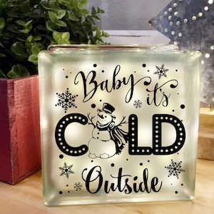 May include: A white glass block with a black silhouette of a snowman and the text "Baby it's COLD Outside".