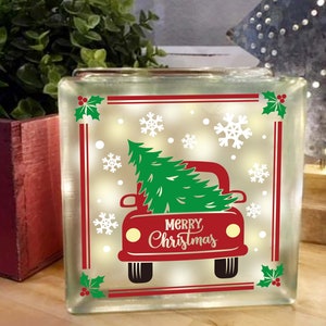 Peut inclure: Un bloc de verre avec un camion rouge transportant un sapin de Noël et les mots "Joyeux Noël". Le camion est décoré de flocons de neige blancs et de houx vert.