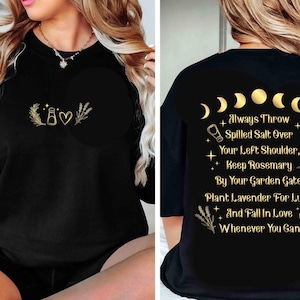 May include: Black sweatshirt with gold-colored graphics. The front features a bottle, heart, and sprig design. The back displays phases of the moon and text: "Always Throw Spilled Salt Over Your Left Shoulder..."